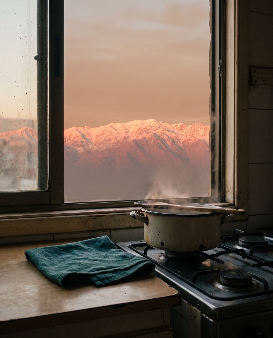 Una ventana de cocina en Santiago al atardecer: la cordillera encendida en rosa sobre la ciudad y, en primer plano, una olla que humea sobre la cocina junto a un paño petrol.