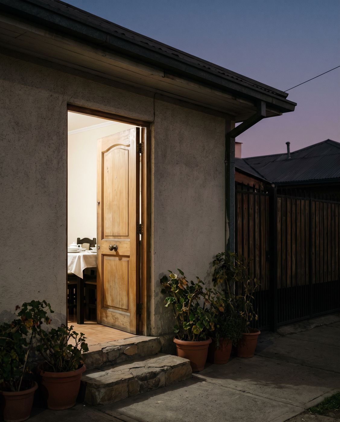 De noche, la puerta de una casa abierta: adentro, luz cálida y el borde de una mesa puesta con mantel blanco; afuera, maceteros de greda junto a la entrada.